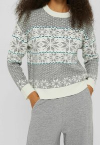 Frau trägt einen grau-weißen Strickpullover mit Schneeflockenmuster und graue Hose, die Hände lässig in den Taschen.