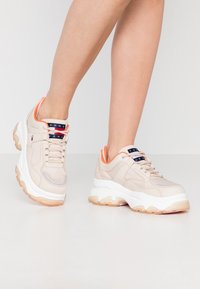 Scarpe sportive beige con suola bianca spessa, tomaia texturizzata, dettagli arancioni e logo sulla linguetta.