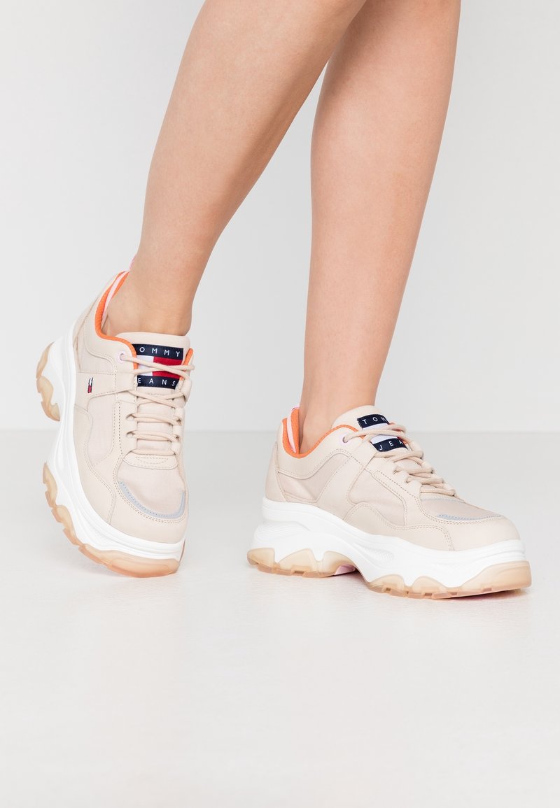 Scarpe sportive beige con suola bianca spessa, tomaia texturizzata, dettagli arancioni e logo sulla linguetta.