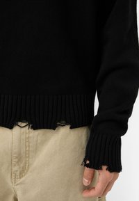 Svart stickad tröja med ribbad nederkant och mudd, med fransiga kanter. Kombinerad med ljusbeige byxor som visar struktur och detaljer.