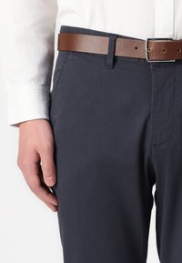 Pantalons texturés bleu marine avec une ceinture en cuir marron. Vue en gros plan mettant en avant la taille et la position de la main sur le côté du pantalon.