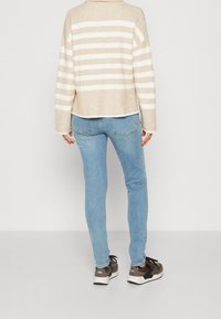 Osoba w beżowo-białym paskowanym swetrze, jasnoniebieskich dżinsach i szarych trampkach, stojąca twarzą do białej ściany.