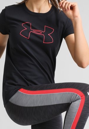 Mujer con camiseta negra con logo rojo de Under Armour y mallas deportivas negras, grises y rojas, realizando un ejercicio de elevación de rodilla.