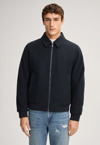 Donker marineblauwe rits bomberjack met een gestructureerde stof, kraag en geribde manchetten. Gepaard met lichtblauwe versleten jeans.