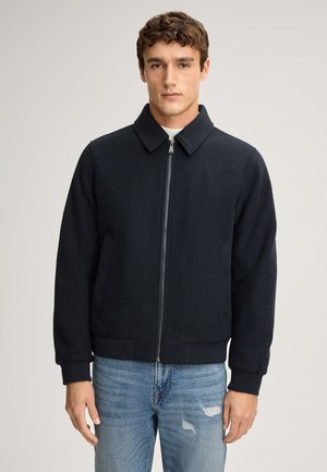 Dunkelblaues, zip-in Bombersakko aus strukturiertem Stoff, mit Kragen und gerippten Manschetten. Kombiniert mit hellblauen, verwaschenen Jeans.