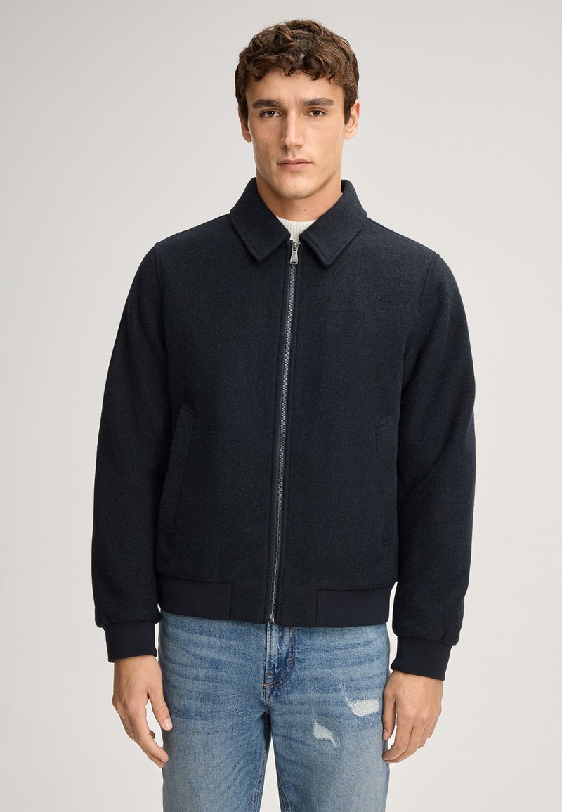 Donker marineblauwe rits bomberjack met een gestructureerde stof, kraag en geribde manchetten. Gepaard met lichtblauwe versleten jeans.