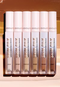 Set med sex concealer-rör med en glänsande rosa finish och klara baser, etiketterade från vänster till höger: Fair Cool, Light Peach, Medium Yellow, Tan Golden, Dark Orange, Deep Red.