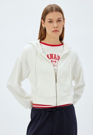 Weißer, taillierter Hoodie mit einem Reißverschluss an der Vorderseite, zwei Taschen und einer Kapuze mit Kordelzug. Über einem weißen Shirt mit rotem Text und blauen Hosen getragen.