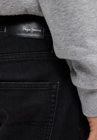 Μαύρα τζιν παντελόνια από denim με ορατές πίσω τσέπες και μια δερμάτινη ετικέτα που γράφει «Pepe Jeans London». Επίσης είναι ορατό γκρι ύφασμα από φούτερ.