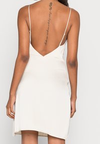 Bílá šatová šaty s hlubokým V-výstřihem na zádech, tenkými ramínky a volnou siluetou, které ukazují hladkou saténovou texturu.