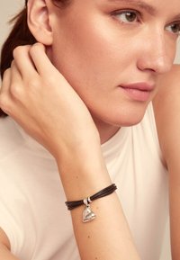 Pulsera de cuero con tres hebras, que cuenta con un charm de corazón plateado grabado con la palabra "UNILTED". El charm aporta un acento reflectante.