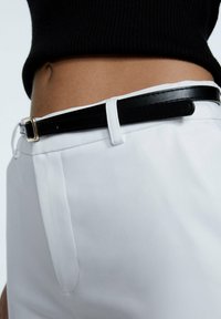 Pantalones blancos con un acabado de tela suave, que presentan un cinturón negro con una hebilla dorada. La cintura tiene un diseño a medida y costuras sutiles.