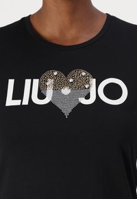 Mustat puuvillapaidat, joissa on valkoinen logo, jossa lukee "LIU JO". Sydämen muotoinen design, jossa on kultaisia ja hopeisia strasseja eri kokoisina. Lyhyet hihat.
