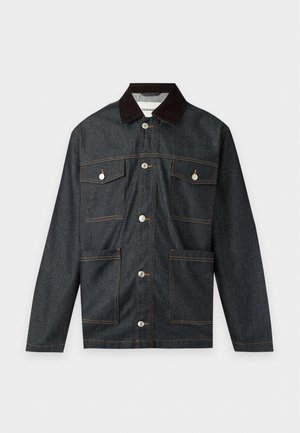 Un homme porte une veste en jean foncée sur une chemise à carreaux verts, un pantalon marron, des bottes noires et une chapka en fourrure foncée, sur un fond uni.