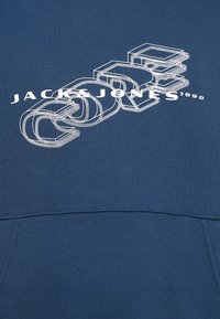 Zils kapucis ar baltu grafisku tekstu, kurā ir "JACK & JONES" un stilizēti burti. Izgatavots no mīkstas, teksturētas auduma.