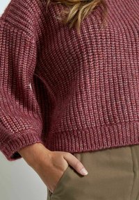 Burgunderfarbener Strickpullover mit geripptem Muster, lockerer Passform und breiten Bündchen. Kombiniert mit olivgrünen Hosen, die Hand in der Tasche ruhend.