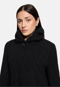 Zwarte fuzzy jas met een grote capuchon, voorzien van een sluiting met knopen aan de voorkant en een gestructureerde stof. Eenvoudig ontwerp, ideaal voor warmte en comfort.