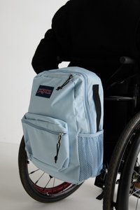 Light blue backpack with a main zip compartment, front pocket, and mesh detail. Features black zippers and straps. Attached to a wheelchair.
