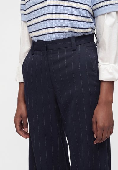 Personne portant un pantalon taille haute bleu marine à fines rayures avec poches latérales et un pull à manches courtes rayé bleu rentré dans une chemise blanche à manches longues.