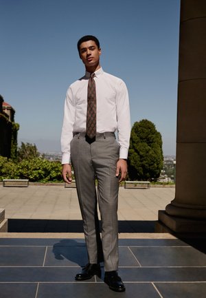 Giovane uomo con pantaloni grigi, camicia bianca, cravatta fantasia e scarpe nere in piedi all'aperto su un patio piastrellato con vegetazione e vista sulla città alle spalle.