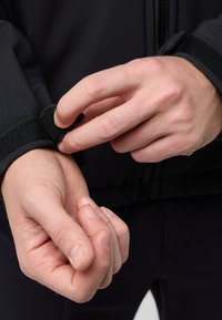 Veste noire au toucher doux, dotée d'une fermeture velcro au niveau du poignet. Des mains ajustent le poignet pour un ajustement confortable.