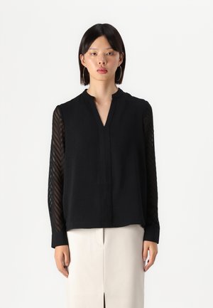 Svart bluse med mandarin-krage, gjennomsiktige ermer med chevron-mønster og avslappet passform. Materialet ser lett og teksturert ut.