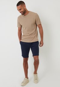 Beige strukturiertes Kurzarmhemd kombiniert mit navyblauen Shorts und hellen Sneakers. Das Outfit hat eine lockere Passform und einen lässigen Stil.