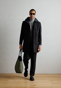Svart frakk over grå hettejakke, svarte bukser og svarte sko. Holder en grønn kviltet totebag. Solbriller fullfører stilen.