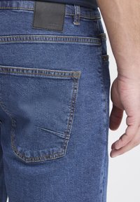 Zilas džinsa bikses ar šauru piegriezumu, redzamām šuvēm un melnu ādas ieliktni pie jostas. Kabatu detaļas ietver nelielu monētu kabatu.