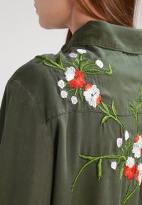 Olivově zelená košile s květinovou výšivkou, na které jsou zřetelné rudé, bílé a purpurové květiny, viditelné stehy a detail límečku na zadní straně.