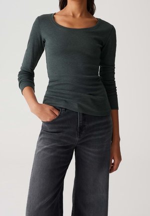 Kobieta w ciemnozielonym topie z długim rękawem i dekoltem w łódkę oraz wyblakłych czarnych dżinsach, stojąca na tle jednolitego tła, z jedną ręką w kieszeni.