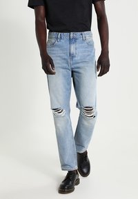 Ljust blå denimjeans med trasiga knädetaljer, rak benpassform och traditionell femfickorsdesign, parat med svarta skor.
