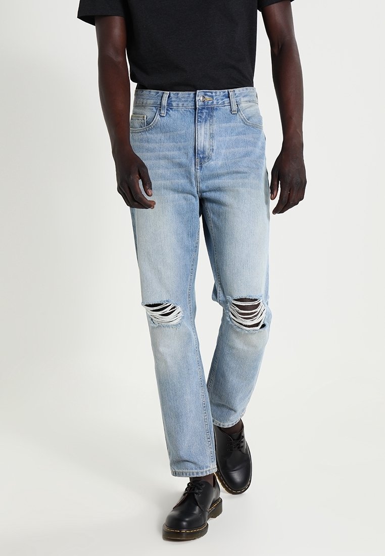 Ljust blå denimjeans med trasiga knädetaljer, rak benpassform och traditionell femfickorsdesign, parat med svarta skor.