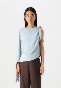 Top satin azzurro chiaro, con una spalla e un dettaglio annodato, texture liscia, vestibilità drappeggiata, abbinato a pantaloni marroni a gamba larga.