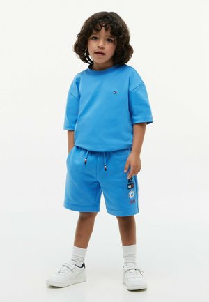 Jeune enfant aux cheveux bouclés portant une chemise bleue à manches courtes et un short assorti avec des baskets blanches et des chaussettes sur un fond uni.