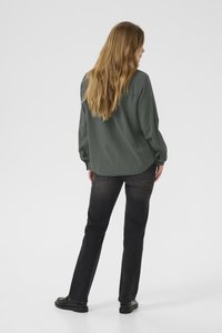 Osoba s dugom svijetlosmeđom kosom koja nosi široku tamnozelenu košulju, crne traperice i crne cipele, stoji okrenuta leđima na bijeloj pozadini.