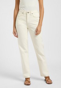 Jeans blancs taille haute en coupe droite, faits de denim doux avec des boutons en métal argenté et des poches avant. Associés à des sandales brunes à lanières.