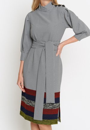 Vrouw draagt een zwart-wit geruite midijurk met driekwartmouwen, een strik in de taille, knopen op de schouders en meerkleurige horizontale strepen bij de zoom.