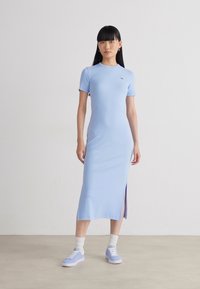 Robe midi ajustée bleu clair avec des manches courtes et une fente sur le côté. Associée à des baskets bleu clair et des chaussettes blanches, avec un petit détail de logo.