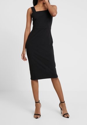 Robe noire ajustée avec un décolleté carré et de larges bretelles. Modèle à longueur genou avec une texture lisse. Associée à des talons noirs à brides.