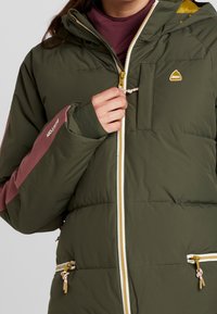 Närbild på en person som bär en huva olivgrön puffy-jacka med rosa detaljer, som har en dragkedja ficka och en guldfärgad dragkedja.