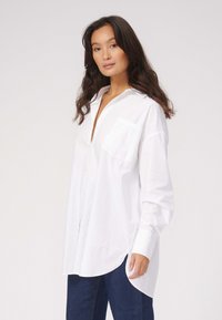 Hvid skjorte med knapper og krave, lomme på venstre bryst og lange ærmer. Stoffet ser glat og en smule struktureret ud.