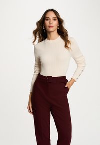 Mujer con cabello castaño ondulado que lleva pendientes de aro dorados, un top de manga larga acanalado en color crema, y pantalones burdeos de tiro alto con un cinturón.