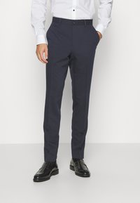 Marineblaue, taillierte Hosen mit flachem Frontdesign, Seitentaschen und schlanker Passform. Kombiniert mit schwarzen Dressschuhen und einem weißen Langarmhemd.