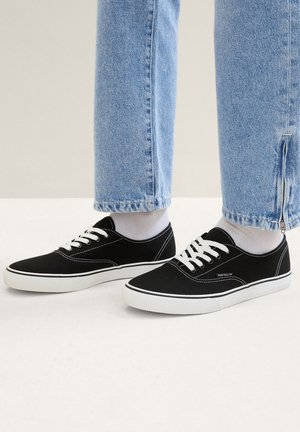 Schwarze Canvas-Sneaker mit weißen Sohlen und Schnürsenkeln. Merkmale eine glatte Textur und ein dezentes Logo-Detail. Kombiniert mit hellblauen Jeans.