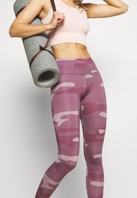 Różowe legginsy w kamuflażu z wysokim stanem, wykonane z elastycznego materiału, w połączeniu z jasnoróżowym topem cropped oraz zwiniętą szaro-żółtą matą do jogi.