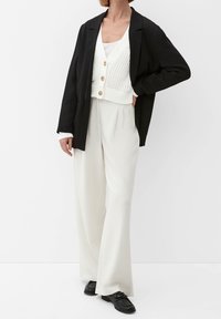 Svart oversized blazer över en vit stickad cardigan med knappar, parat med lösa vita byxor och svarta skor. Minimalistisk design och färger.