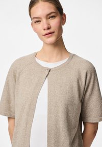 Cardigã bege claro com mangas curtas, textura suave e decote redondo. Apresenta um fecho com um único botão na frente.