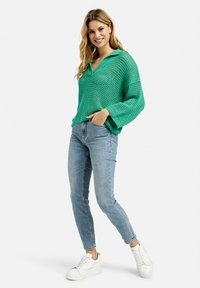 Grüner, offener Strickpullover mit Kragen, kombiniert mit hellblauen Jeans und weißen Sneakers. Das Outfit hat eine lockere Passform und einen lässigen Stil.