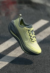 Αθλητικό παπούτσι Nike ZoomX σε φωτεινό κίτρινο χρώμα, με υφασμάτινο επάνω μέρος, γκρι λεπτομέρειες και απορροφητική σόλα, κρεμασμένο πάνω από επιφάνεια από σκυρόδεμα.